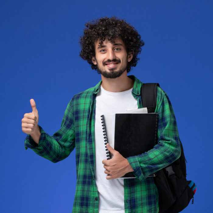 front-view-male-student-green-checkered-shirt-with-black-backpack-holding-copybooks-smiling-blue-wall