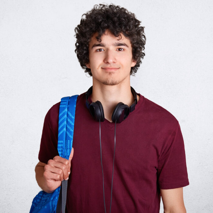 Portrait of delighted hipster male student with crisp hair, wears casual t shirt, carries backpack, has headphones on neck, ready to go for classes at university, isolated over white background