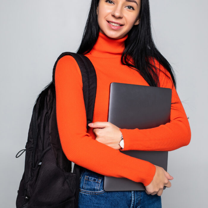 young-student-girl-isolated-gray-wall-smiling-camera-pressing-laptop-chest-wearing-backpack-ready-go-studies-start-new-project-suggest-new-ideas